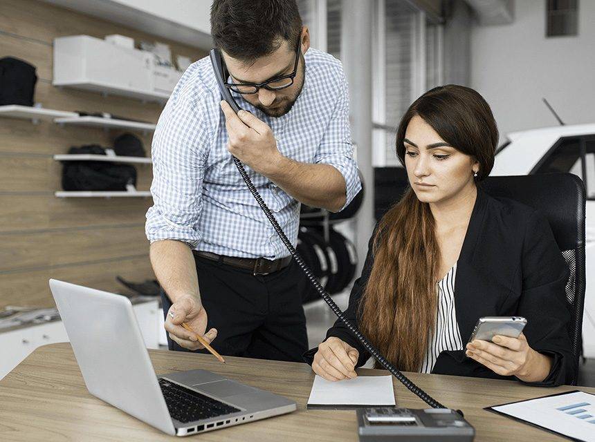 Dois profissionais em um ambiente de escritório. O homem, de óculos e camisa xadrez, está ao telefone enquanto aponta para o laptop com um lápis. A mulher, ao lado dele, está concentrada, segurando um smartphone e tomando notas em um bloco de papel. Ambos parecem estar colaborando em uma tarefa, com o ambiente sugerindo uma atmosfera de trabalho produtiva e focada.